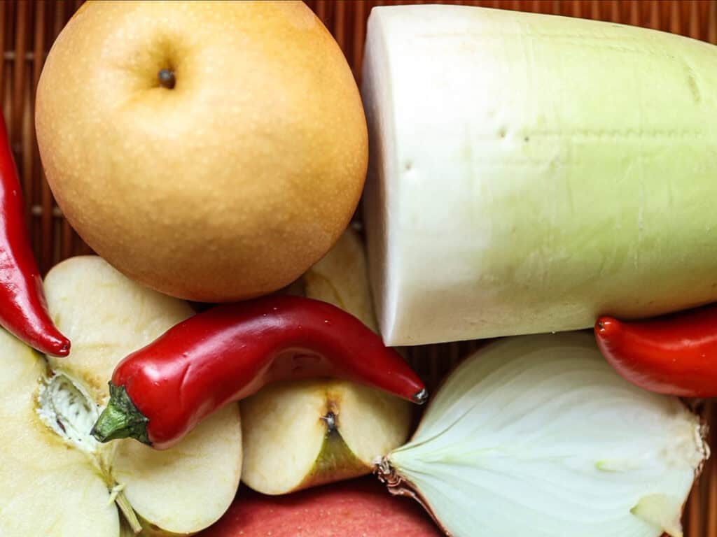 A variety of fresh produce including a whole Asian pear, daikon radish, sliced apples, two red chili peppers, and a cut onion displayed on a wooden surface.